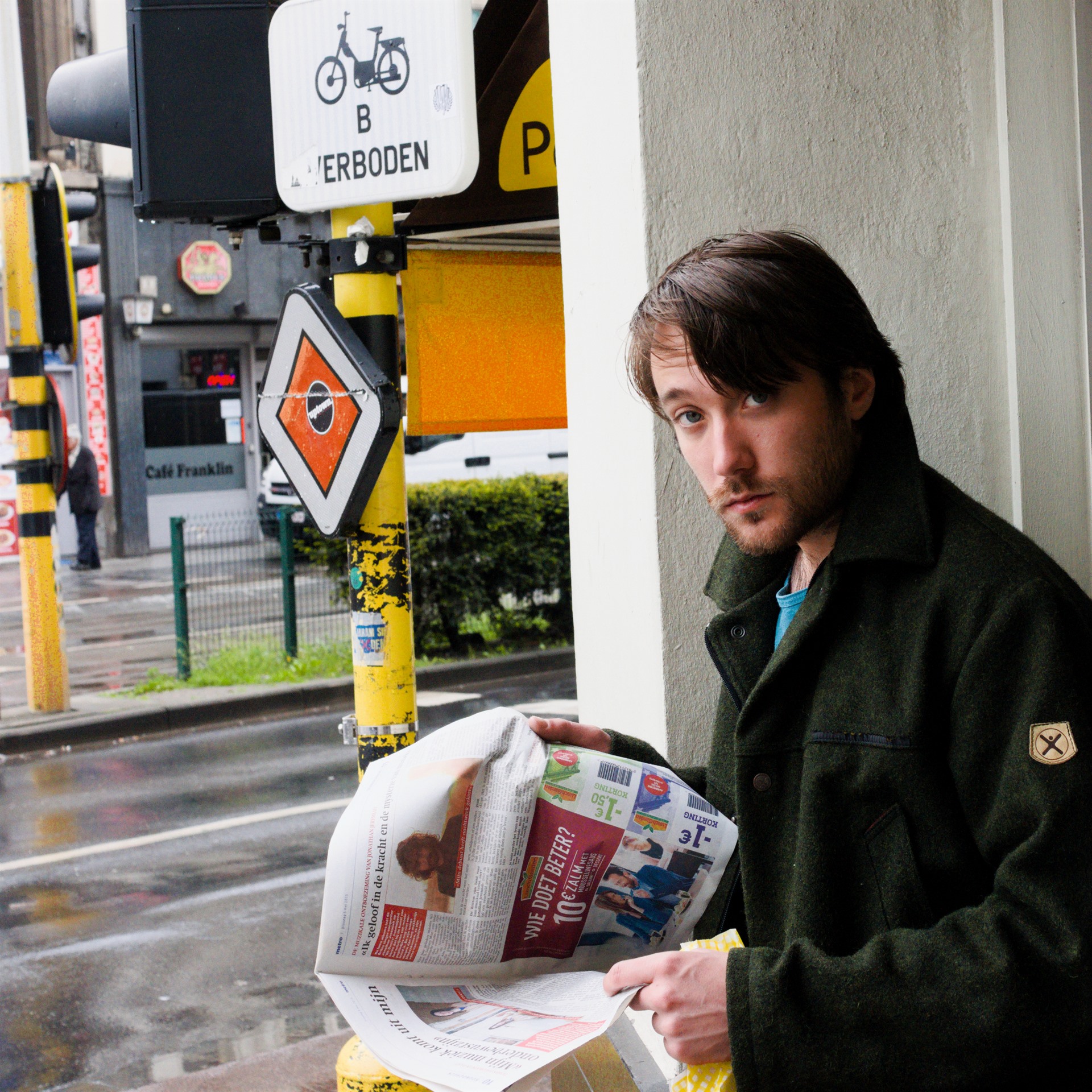 Man holding a newspaper beside a window overlooking traffic signs and a wet street.