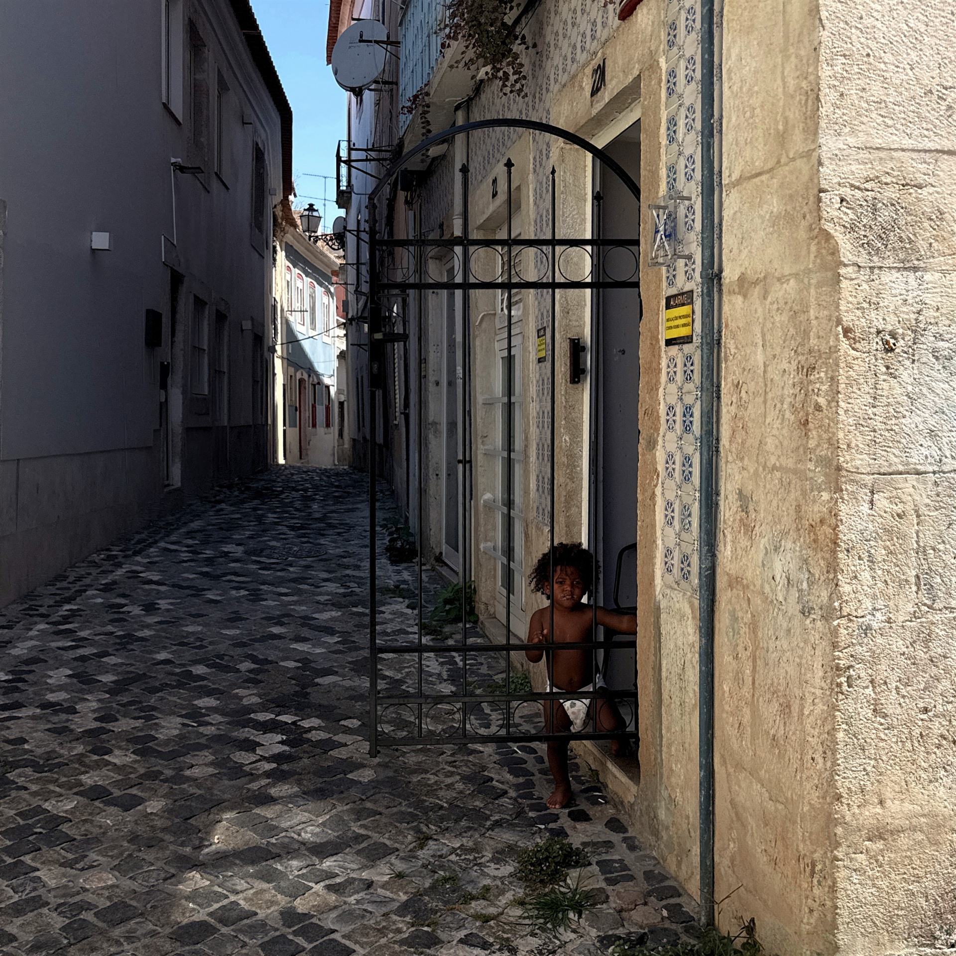 Child standing behind an iron gate in a narrow tiled alley.