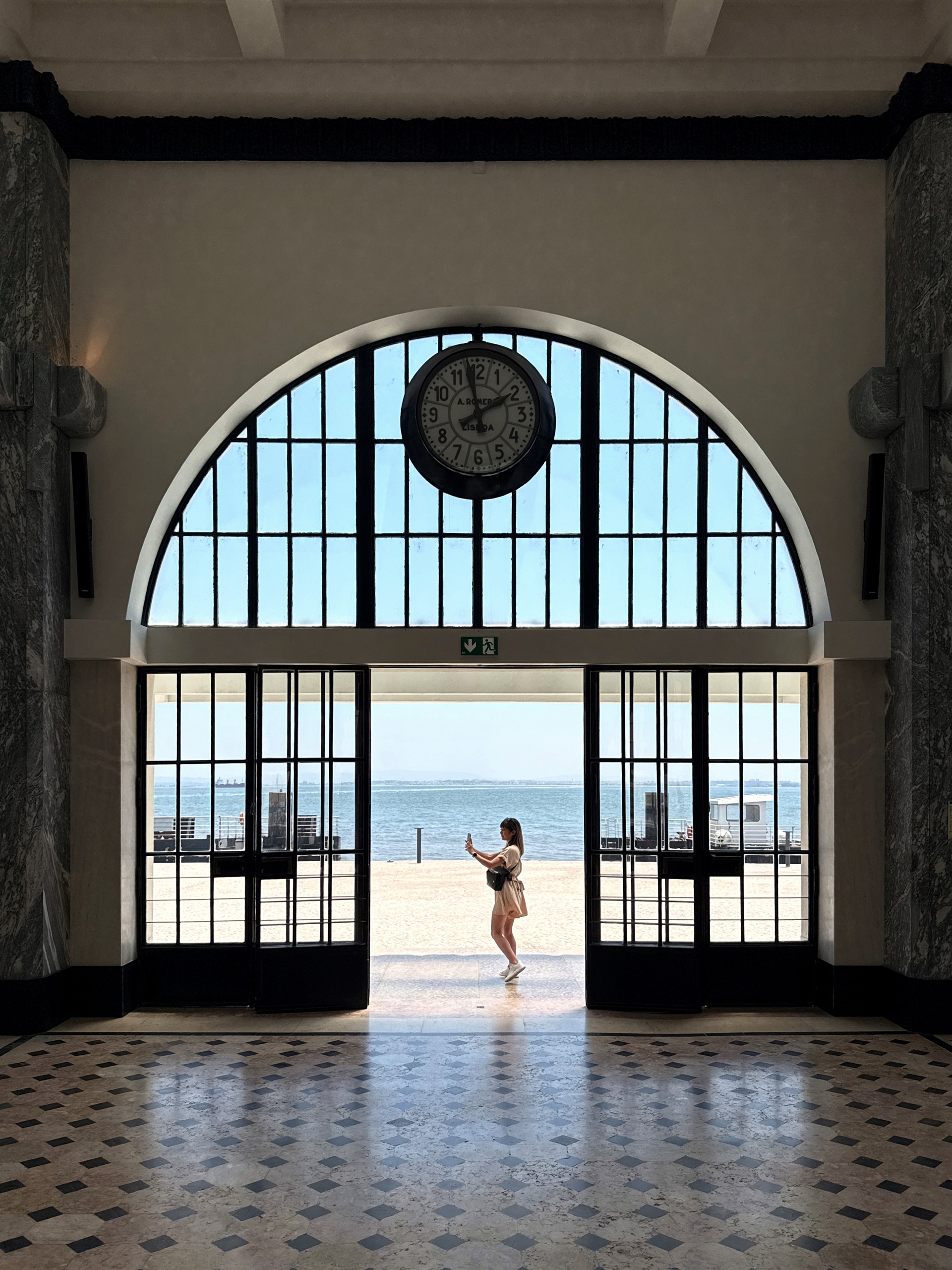 Woman standing beneath a large clock in a monumental seaside hall.
