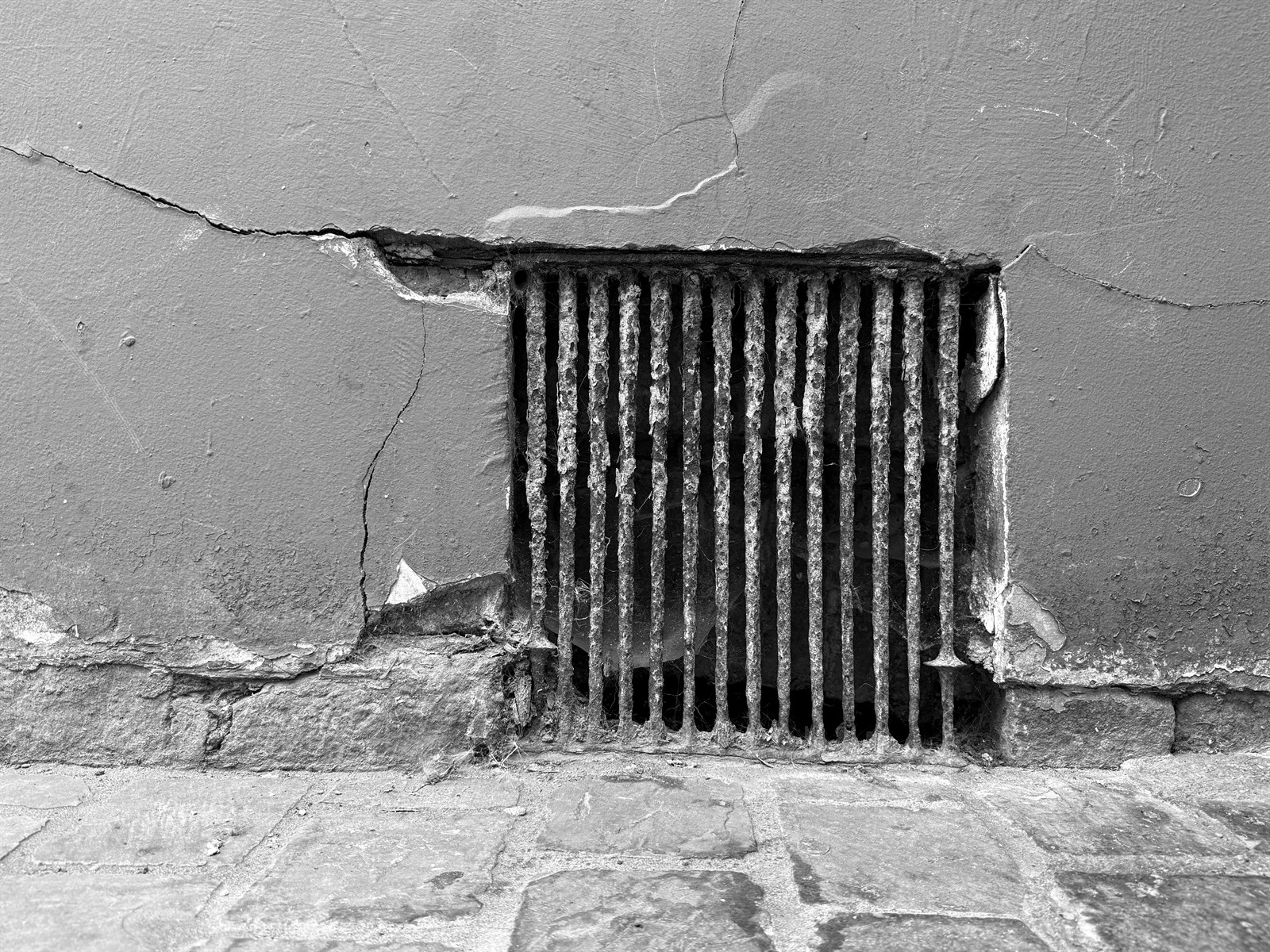 Metal ventilation grille surrounded by cracked and patched render.